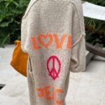 Strikket cardigan med LOVE og PEACE-design i naturmiljø og sennepsgul håndveske.