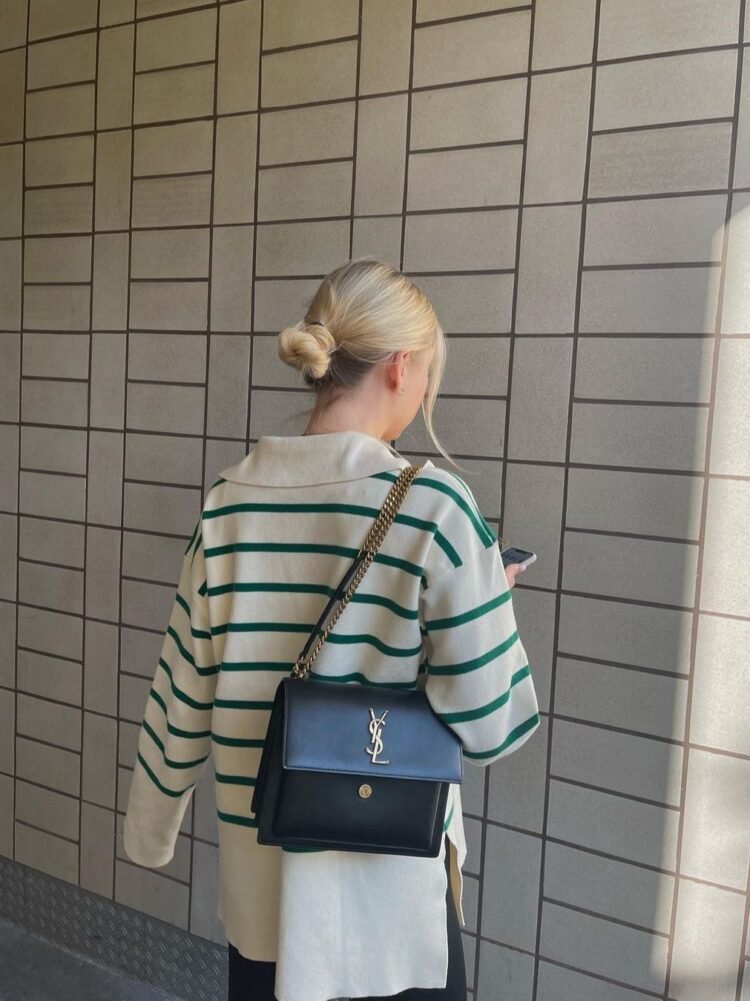 Avslappet person med blond hår og skulderbag ser på mobiltelefon ved flisvegg.