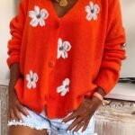 Orange cardigan med hvite blomster, blå denim shorts og gull halskjede på rustikt møbel.